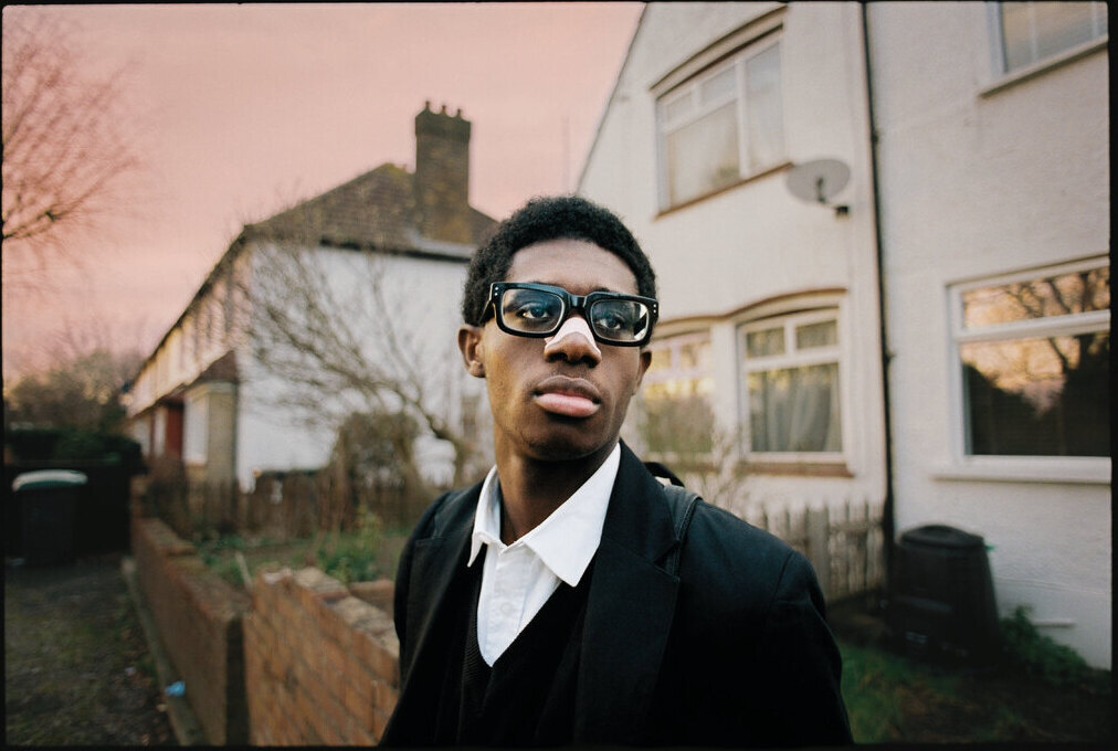A young man in sunglasses outside terraced houses, by Seán Anthony