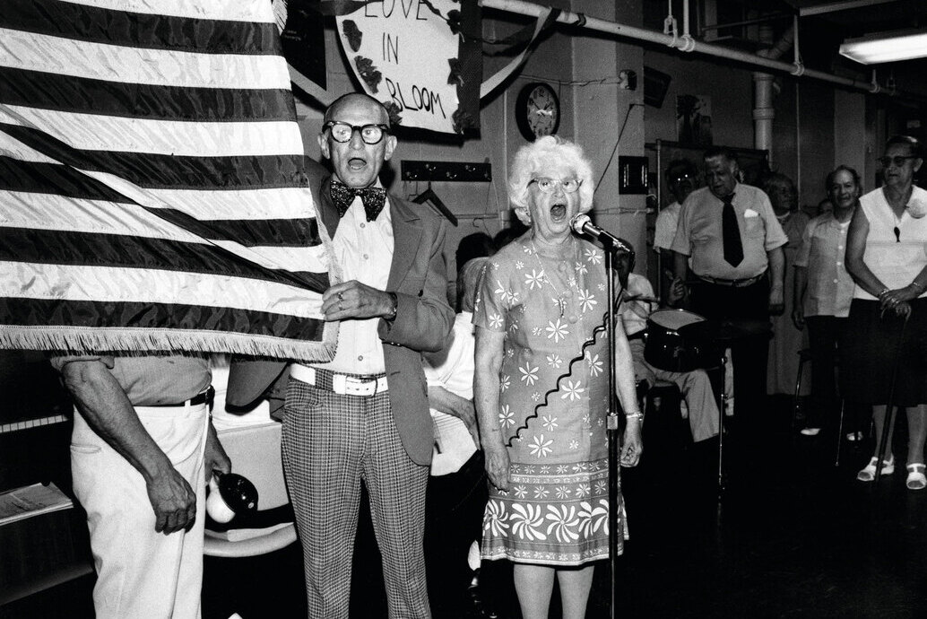 An elderly couple singing beside an American flag, by Meryl Meisler